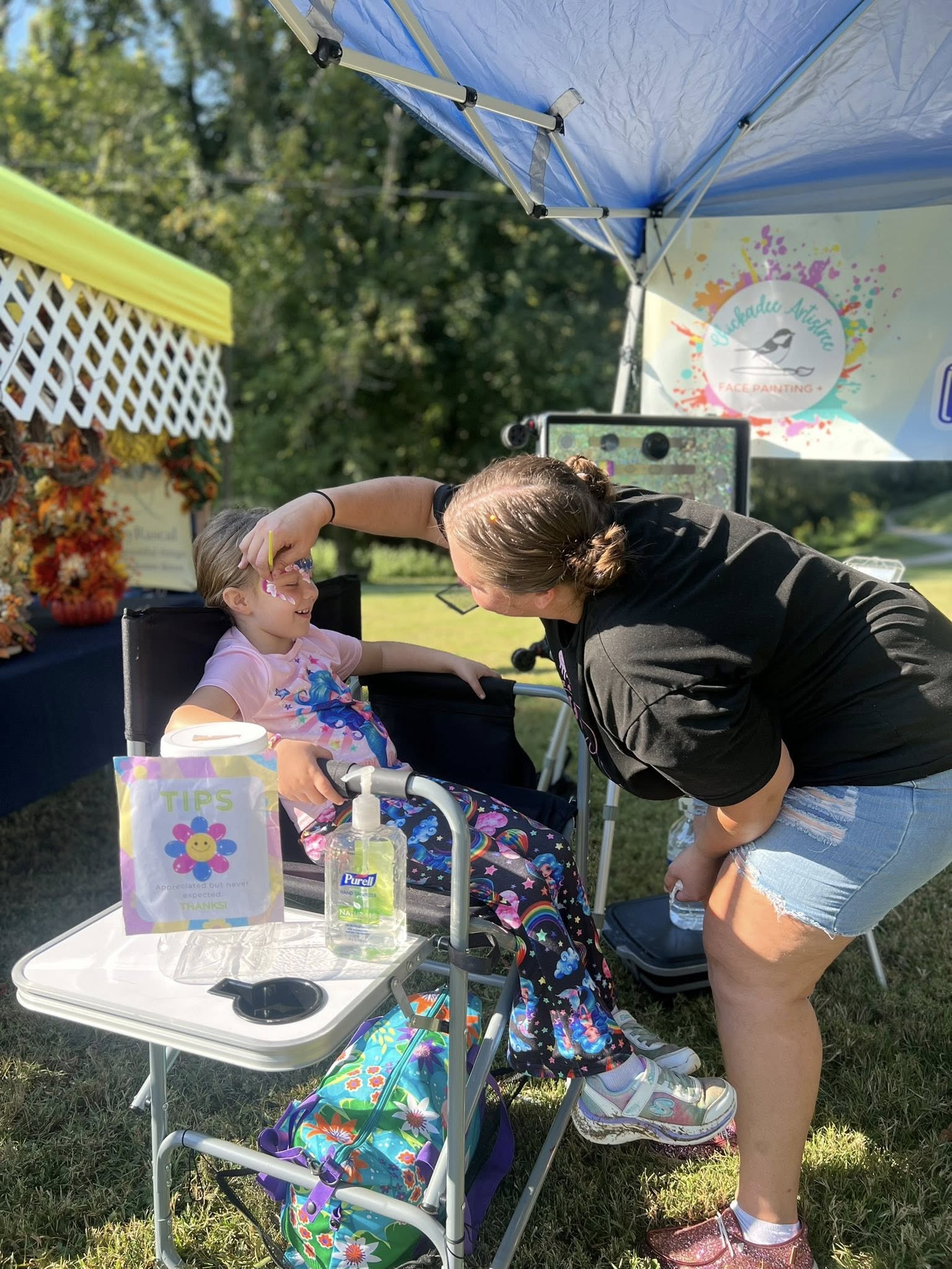 Woman applying face paint to a child's face under a blue canopy tent.