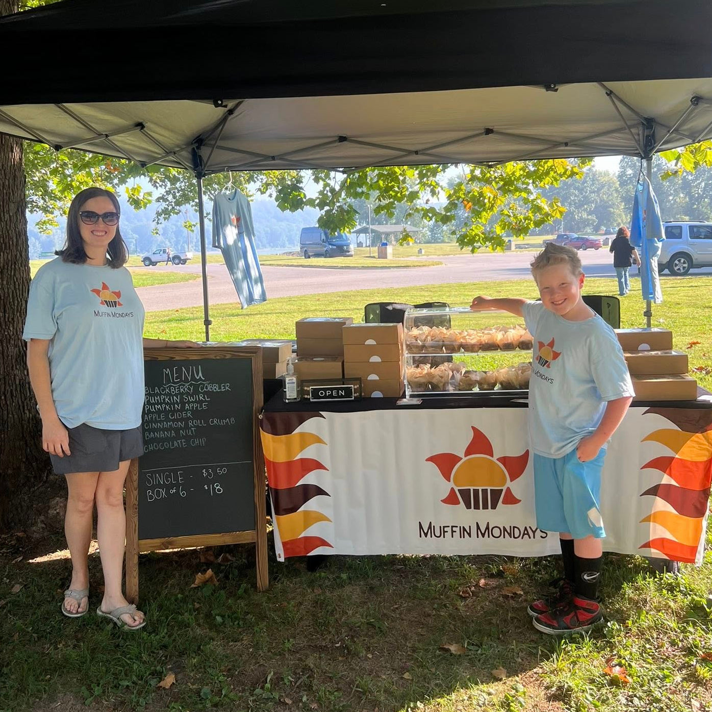 Two people standing under a black canopy with a 'Muffin Mondays' tablecloth at an outdoor event.